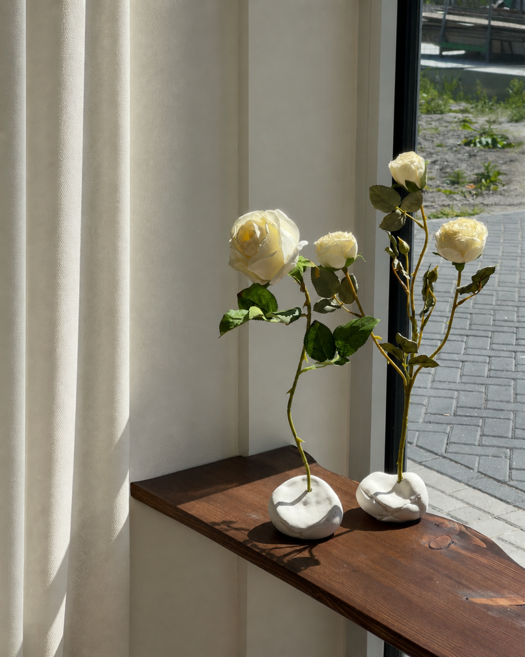 minimalist interior, white roses, ceramic vases, handmade decor, pebble vases, sculptural objects, wooden shelf, window light, sunlight shadows, neutral tones, aesthetic decor, soft natural light, floral arrangement, modern home, calm atmosphere, windowsill styling, organic shapes, artisanal pottery, natural materials, simple living, serene space, cozy corner, contemporary design, clean aesthetic, still life, home styling